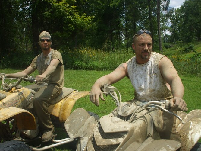 Two muddy riders on ATVs in a grassy yard; foreground quad caked in dried mud, yellow ATV behind