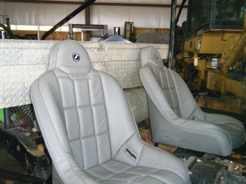 1978 Ford Bronco - Two grey racing seats with headrests are visible in a workshop setting, with a large yellow piece of machinery in the background.