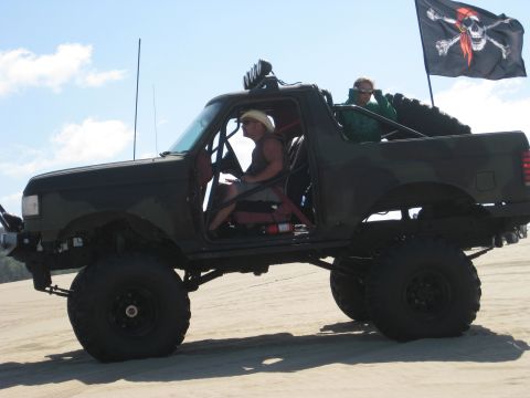 1979 Ford Bronco - A heavily modified SUV with two occupants drives across a sandy terrain under a blue sky.