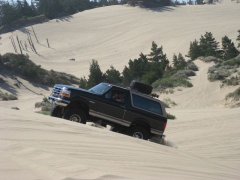 1979 Ford Bronco - Black two-tone lifted Ford Bronco cresting a sand dune with the driver visible through the open window, kicking up sand at the rear tires.…