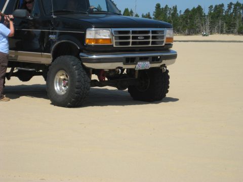 1979 Ford Bronco - This lifted black Ford Bronco is parked on a sandy area with trees in the background.