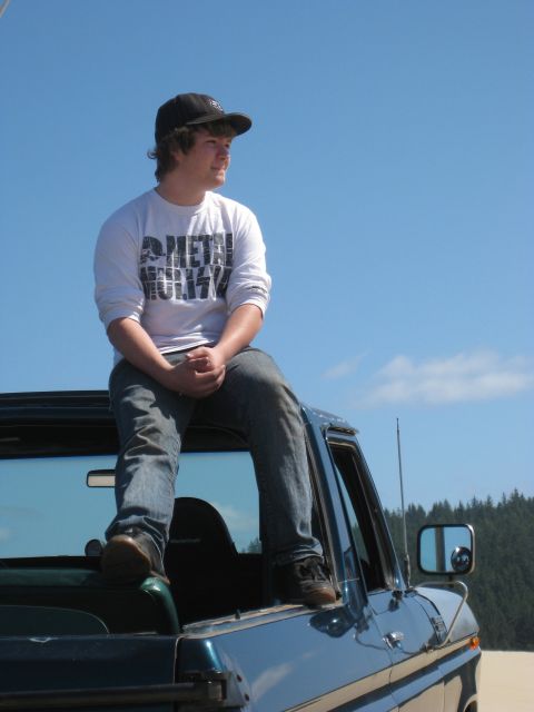 1979 Ford Bronco - A young man in a ball cap and white Metal Militia long-sleeve shirt sits on the open roll bar/roof line of a dark teal-green full-size Ford…