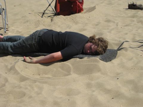1979 Ford Bronco - A person in jeans and a dark T-shirt lies face-down asleep in the sand at a dune campsite, next to a folding chair and a red cooler or bag.…