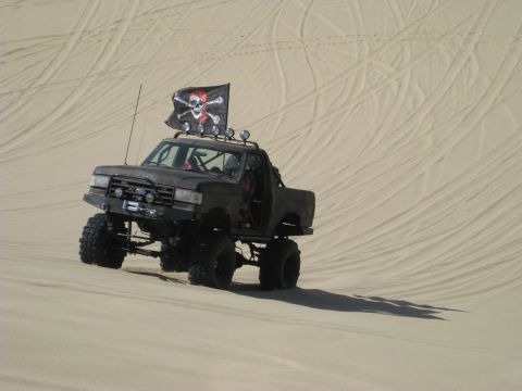 1979 Ford Bronco - Lifted flat-black Ford 4x4 truck with a chopped/topless cab sitting on a sand dune covered in tire tracks, seen from a three-quarter front…