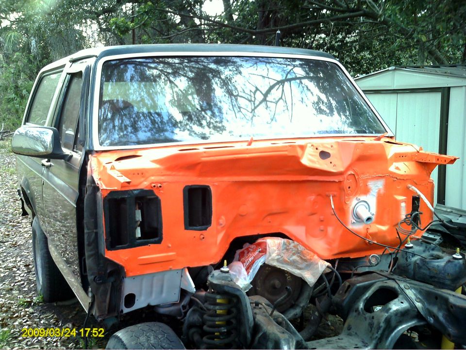Ford Bronco in disrepair, front end disassembled and painted orange. Side view shows dark body panels and chrome mirror