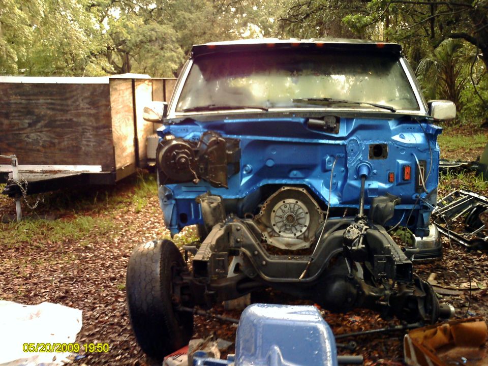 Vehicle chassis with blue painted firewall, transmission housing visible, front wheel attached. Trailer in background
