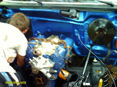 1992 Ford Bronco - A person works on a V8 engine inside a blue-painted engine bay, with a black brake booster visible.