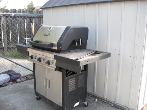 1991 Ford Bronco - A gas grill sits on a patio, with a chain-link fence and shed in the background.