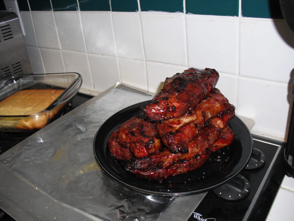 pile of glazed ribs on a black plate next to a pan of cornbread on a stovetop