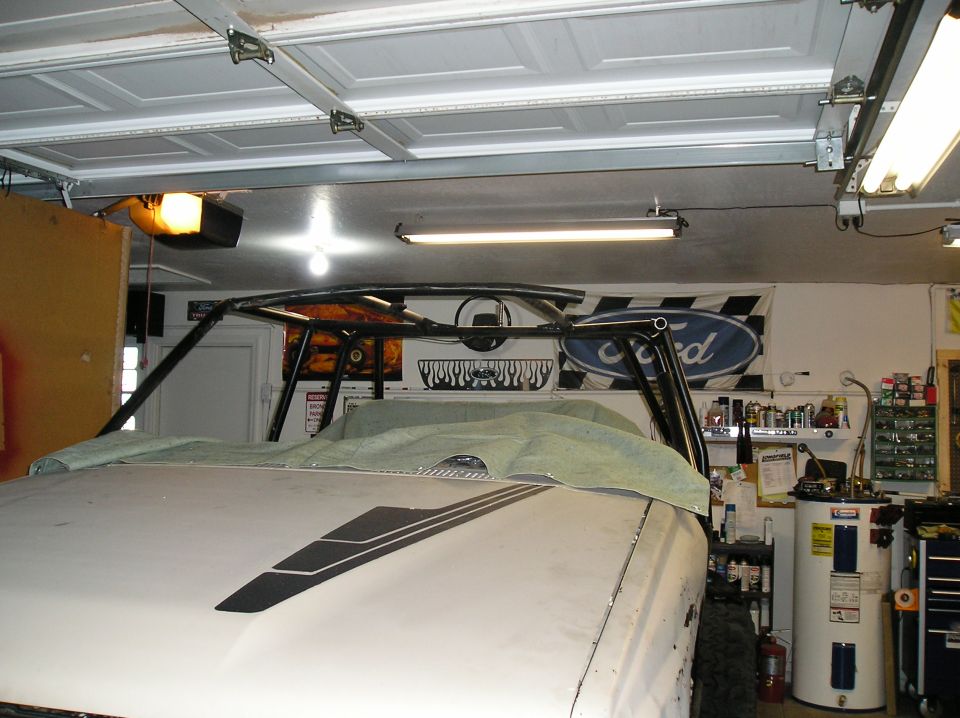 White vehicle hood with black stripes, covered with a tarp, showing a roll cage structure inside a garage. Ford flag visible