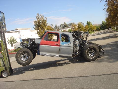 1986 Ford Bronco - A heavily modified truck, likely from the 1970s, is being lifted by a forklift, revealing its custom suspension and roll cage.
