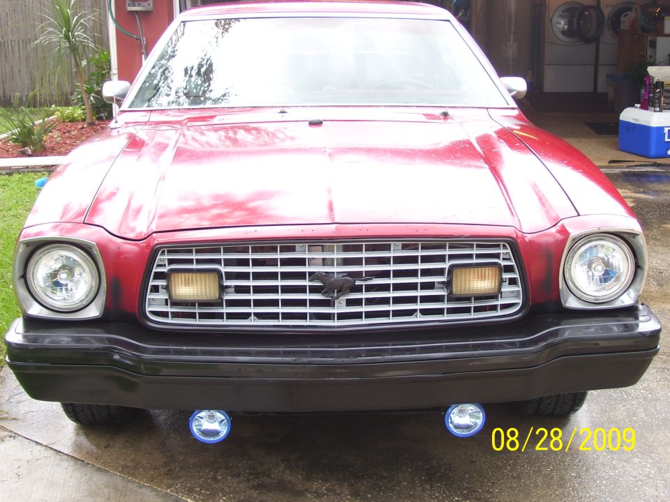 Front of a red Ford Mustang II in a garage with fog lights on