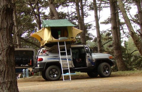 1989 Ford Bronco - An SUV is parked in a wooded area with a rooftop tent deployed, ready for camping.