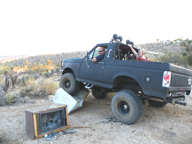 Black lifted Ford Bronco with light bar climbing over an old TV in the desert, rear 3/4 view