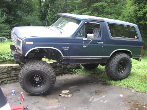 1986 Ford Bronco - A lifted blue Ford Bronco sits on a paved surface with grass and trees in the background.