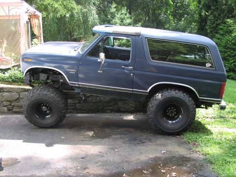 1986 Ford Bronco - A lifted blue Ford Bronco with large tires sits on a paved surface next to a stone wall and grass.