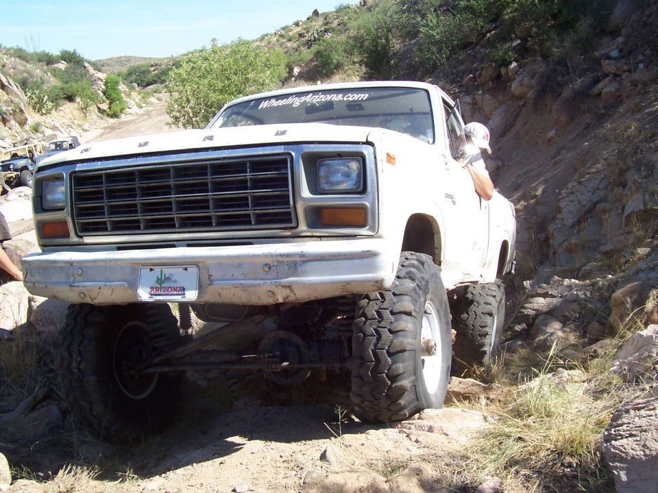 White Ford pickup truck with large tires navigating a rocky trail. Another vehicle is visible in the background