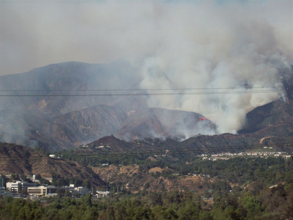 Wildfire burning on a mountainside with smoke billowing. An air tanker is visible dropping retardant