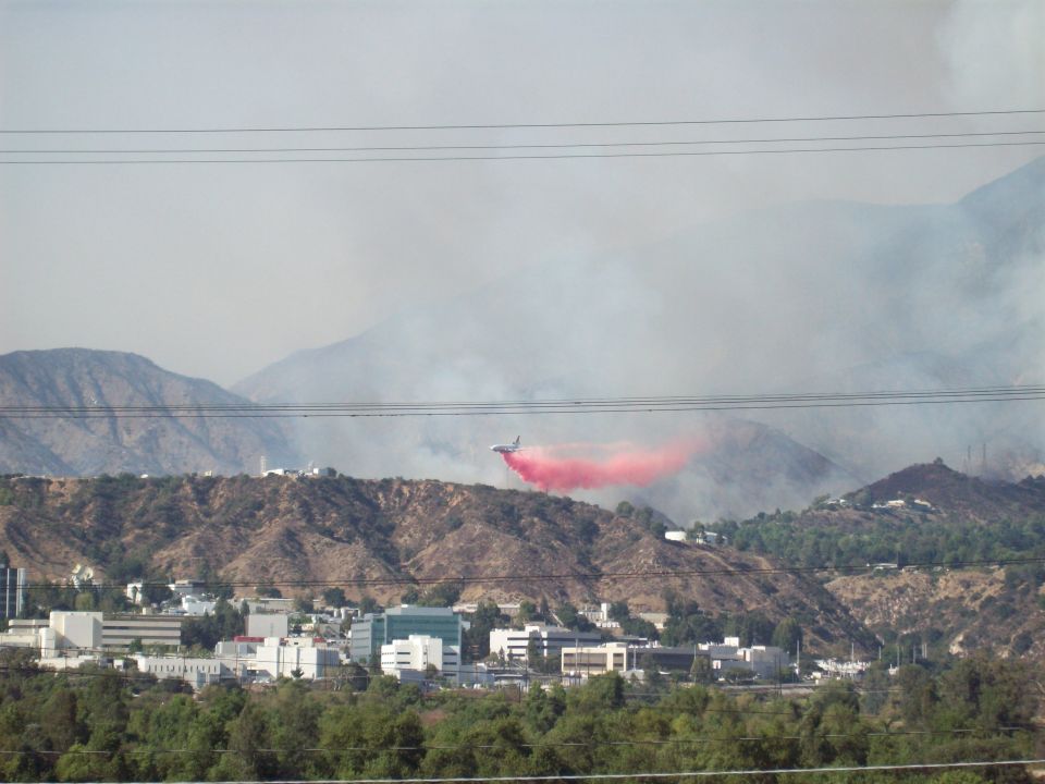 Firefighting aircraft drops red retardant over a hillside with buildings below