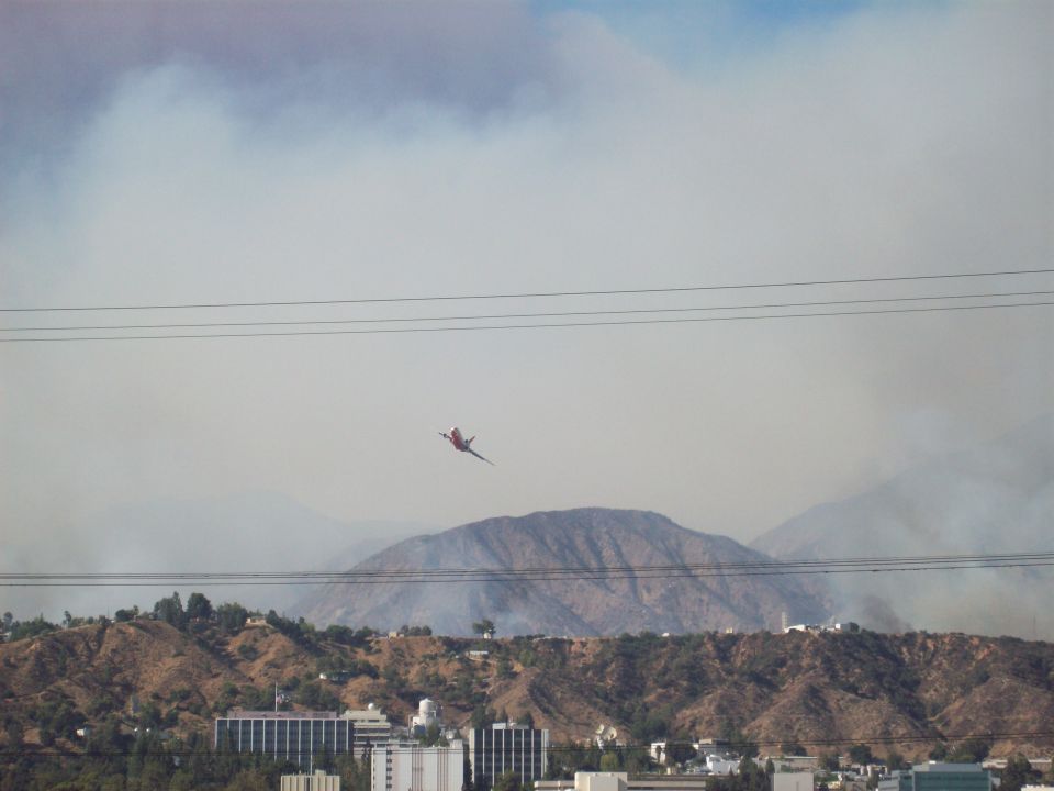 Air tanker flying over hills with smoke and buildings below