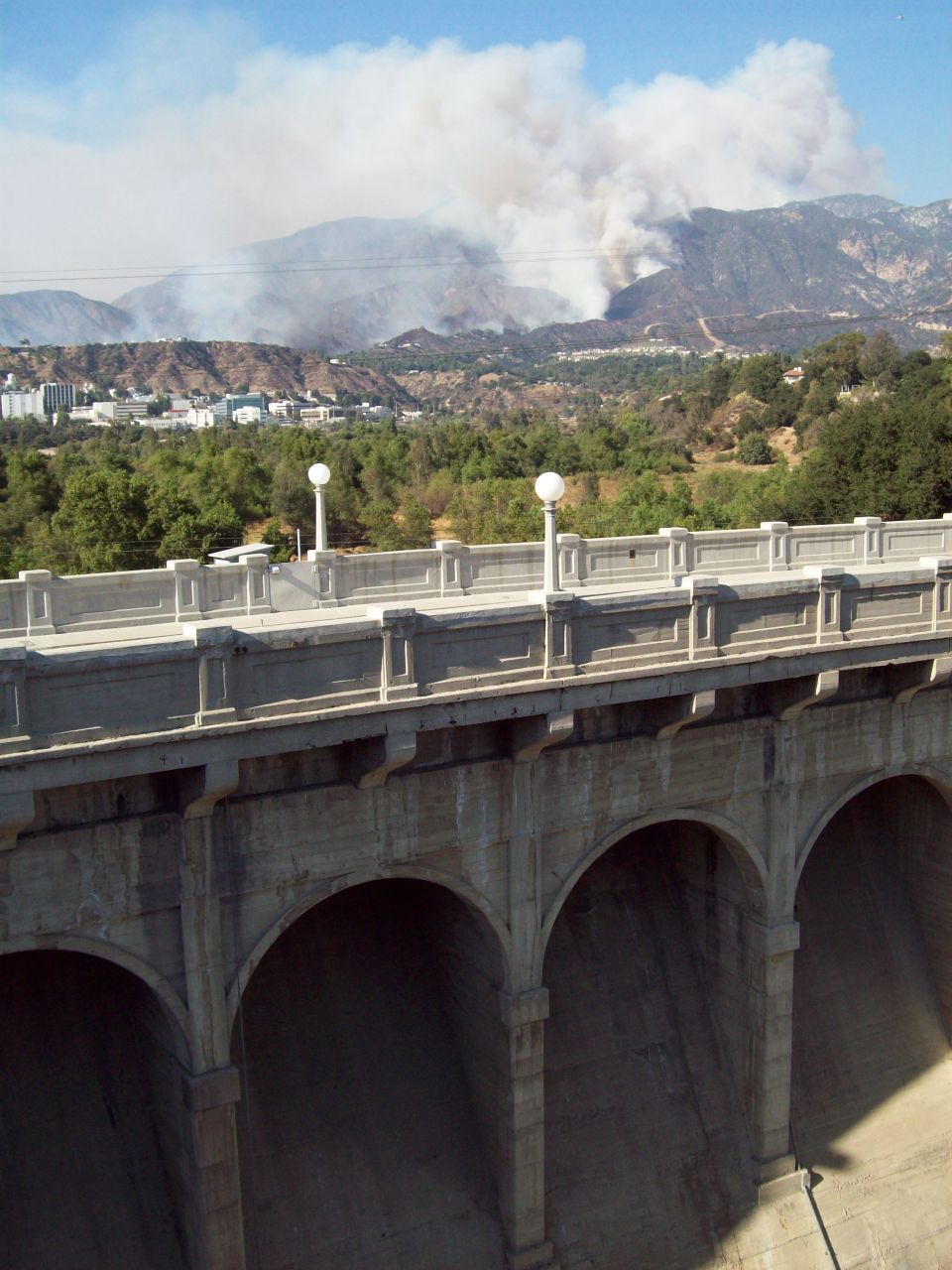 Smoke billows from a wildfire on a distant hillside, viewed over a concrete bridge