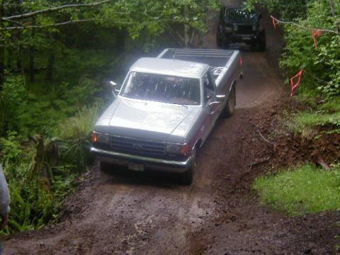 1989 Ford F-150 - A silver pickup truck navigates a dirt path in a wooded area, followed by a dark SUV.