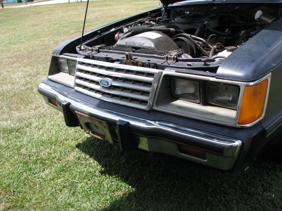 Dark blue Ford car front end with hood open, showing engine. Chrome grille with Ford logo, rectangular headlights