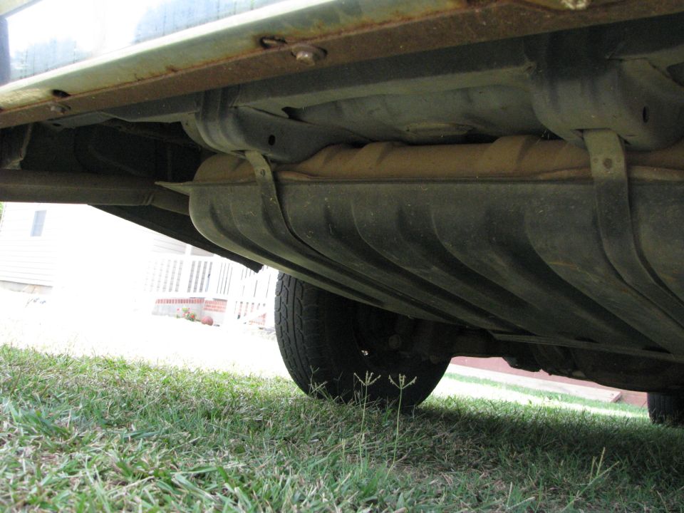 Underside of a car on grass showing ribbed fuel tank, exhaust pipe and rusty rocker seam
