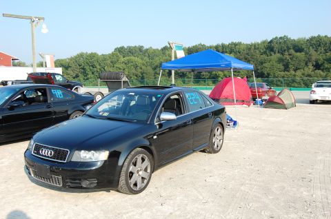 2002 BMW M3 - An Audi sedan is parked at a race track event with tents and other vehicles in the background.