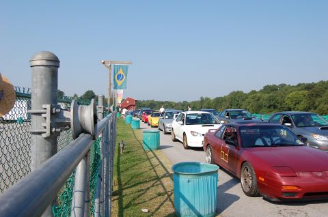 2002 BMW M3 - A line of cars waits on the track under a clear blue sky, with a Brazilian flag flying nearby.