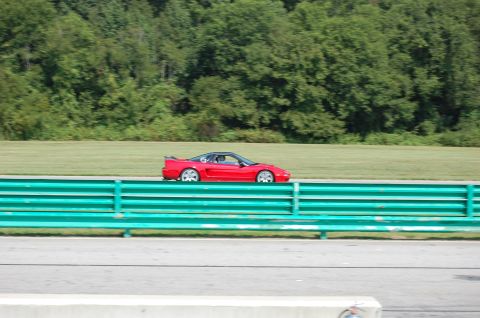 2002 BMW M3 - A red sports car is seen driving on a race track with a green barrier in the foreground.