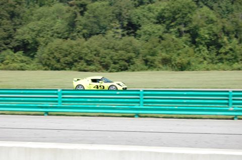 2002 BMW M3 - A yellow race car numbered 49 drives on a track with a green barrier in the foreground.