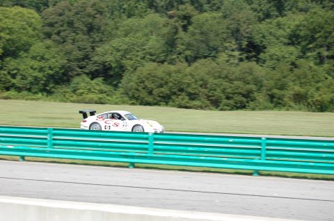 2002 BMW M3 - A white Porsche 911 race car is seen on a track with a green guardrail in the foreground.