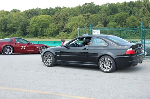 2002 BMW M3 - A black BMW M3 and a red Chevrolet Corvette are on a racetrack with trees in the background.