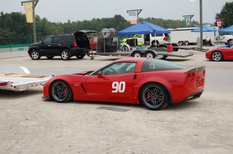 2002 BMW M3 - A red Chevrolet Corvette with racing number 90 is parked at a track event with other vehicles and trailers.