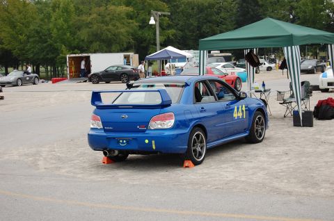 2002 BMW M3 - A blue Subaru WRX STI is parked at a track event with other vehicles and tents in the background.