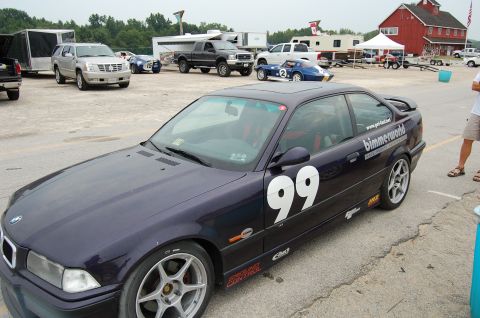 2002 BMW M3 - A BMW coupe with racing decals and number 99 is parked at an outdoor event with other vehicles and structures visible.