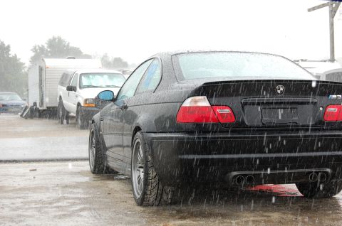 2002 BMW M3 - A black 2002 BMW M3 coupe is seen from the rear in the rain, with other vehicles and a trailer in the background.