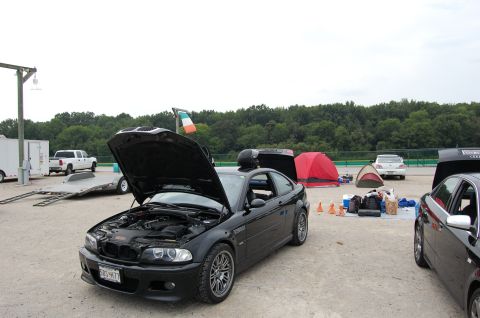 2002 BMW M3 - A black BMW M3 is parked with its hood open, alongside tents and other vehicles at what appears to be a track event.