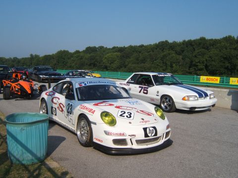 2002 BMW M3 - Two race cars, a Porsche and a Ford Mustang, are parked on a racetrack with trees in the background.