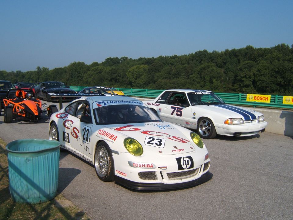White Porsche 911 GT3 Cup car with yellow headlights and a white Ford Mustang with blue stripes on a racetrack