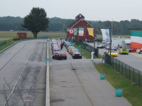 2002 BMW M3 - Cars lined up at a race track with a barn and flags visible in the background.