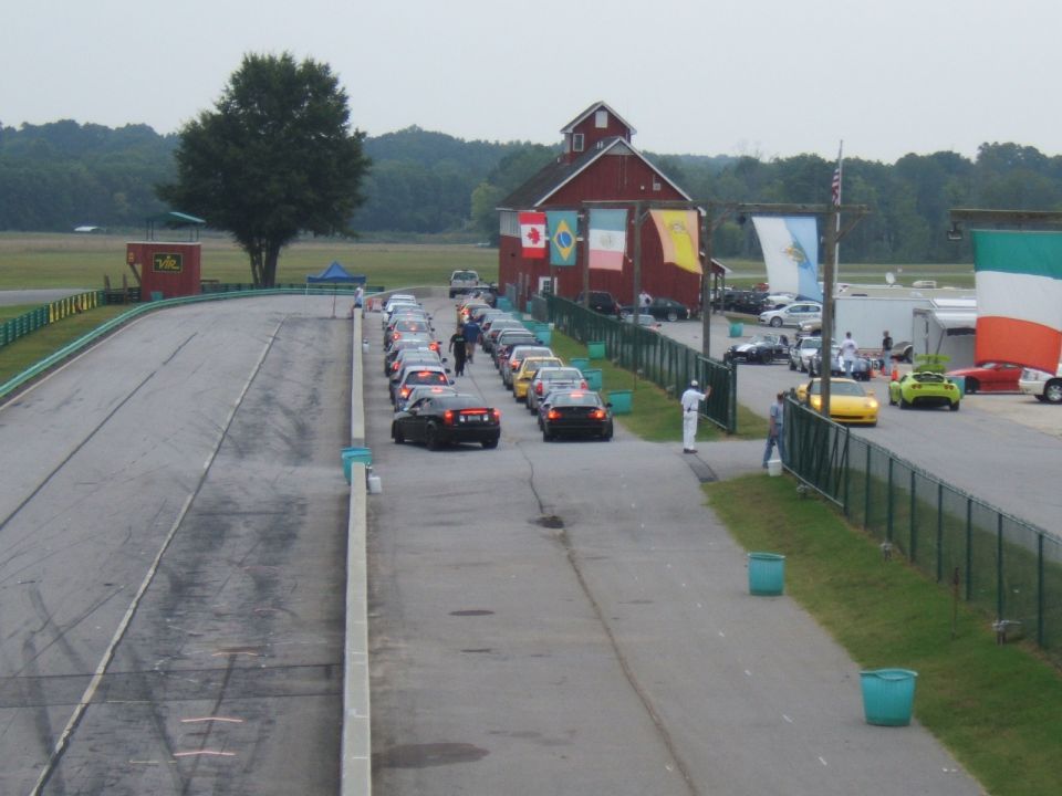 Race track with a line of cars parked on the side, a red barn in the background, and flags hanging