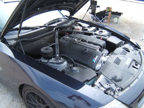 2002 BMW M3 - The engine compartment of a dark blue BMW is shown with the hood open, revealing the engine and various components.