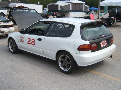 2002 BMW M3 - A white hatchback with racing number 28 and a roll cage is parked at an event with other vehicles and campers in the background.