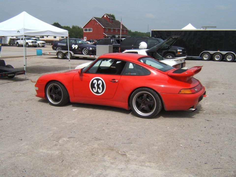 Red Porsche 911 with racing number 33 on door, parked on asphalt with tents and buildings in background