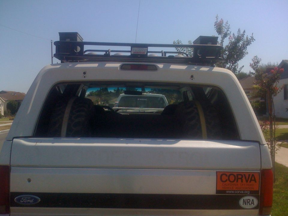 Rear view of white SUV with roof rack and spare tires inside. Another vehicle is visible through the rear window