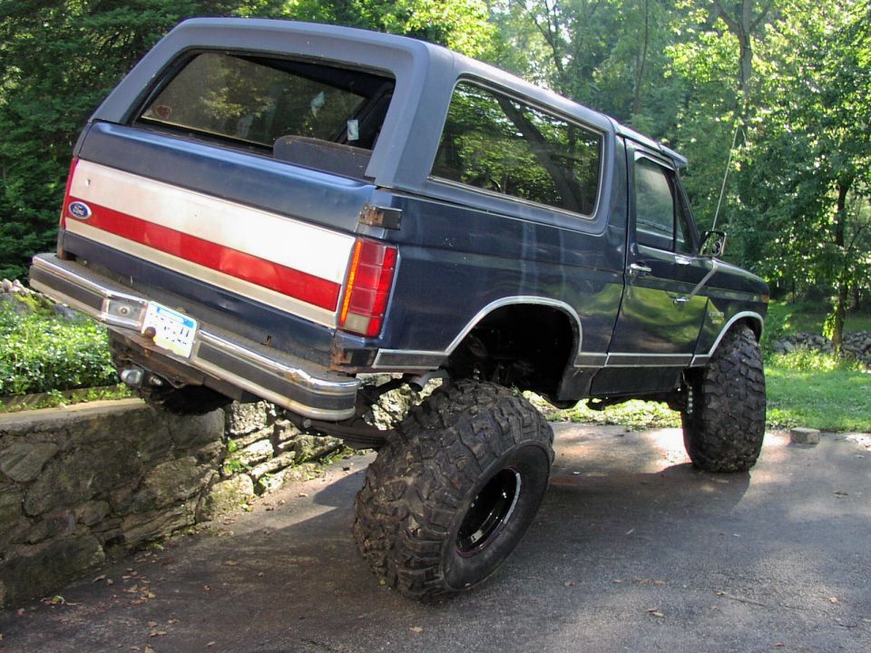 Dark blue Ford Bronco with red and white stripe on the side, lifted with big off-road tires, partially on a stone wall