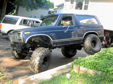 1986 Ford Bronco - A lifted blue 1986 Ford Bronco sits parked next to a white SUV.