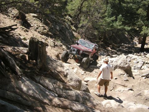 1995 Ford Bronco - A red Jeep Wrangler tackles a challenging off-road course on a sunny day, with a spectator observing the difficult ascent.